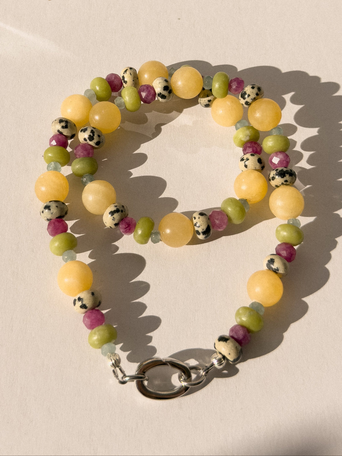 A beaded necklace with a silver clasp, featuring round beads in pale yellow, light green, magenta pink, and speckled black-and-white agate, arranged on a warm beige surface with strong sunlight and shadows.