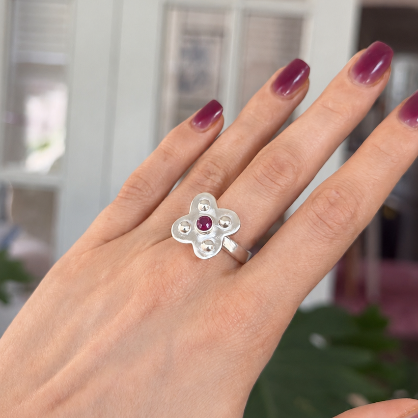 A silver flower-shaped ring with a small red gemstone in the center, worn on a hand with dark purple manicured nails.
